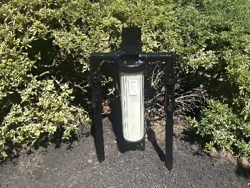A water filtration system with a visible filter cartridge installed on a black frame outdoors in front of a green bush.