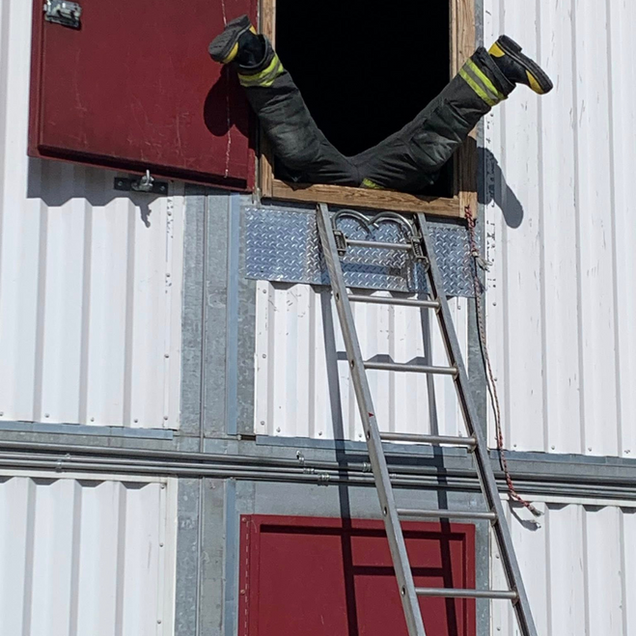 Firefighter in black gear exiting a window, legs first, above a ladder on a white building with red doors.