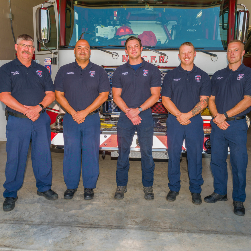 Five firefighters in navy uniforms stand in front of a fire truck, smiling.