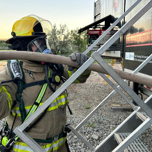 Firefighter in full gear holding a hose near a training trailer. Yellow helmet, neutral expression.