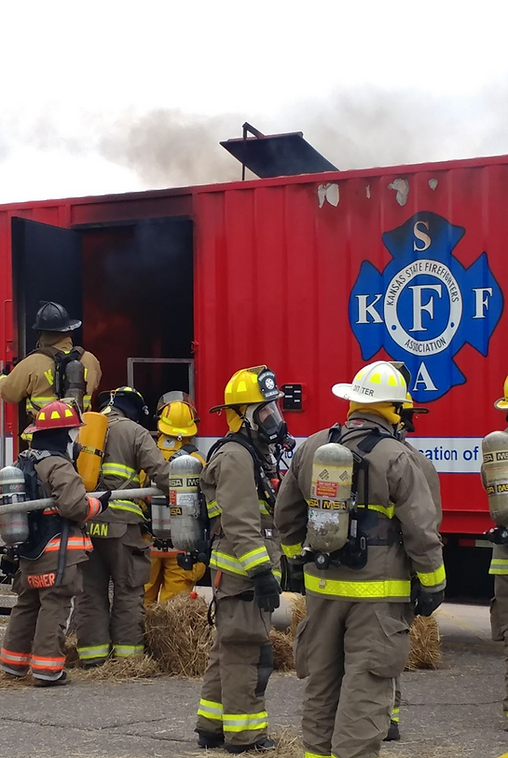 Firefighters in protective gear at a red training structure with smoke billowing out.