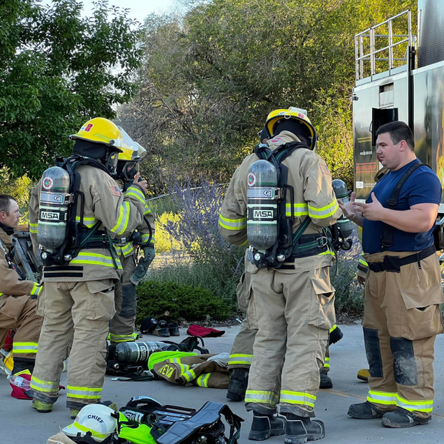 Firefighters in gear standing by truck, taking a break, with equipment on the ground.