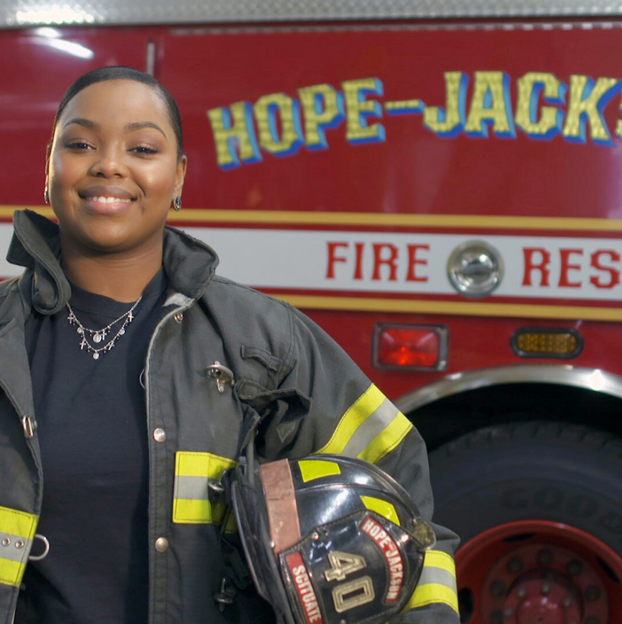 Firefighter smiles in front of a fire truck. She wears a uniform and holds a helmet. Truck reads “Hope-Jackson Fire Rescue.”
