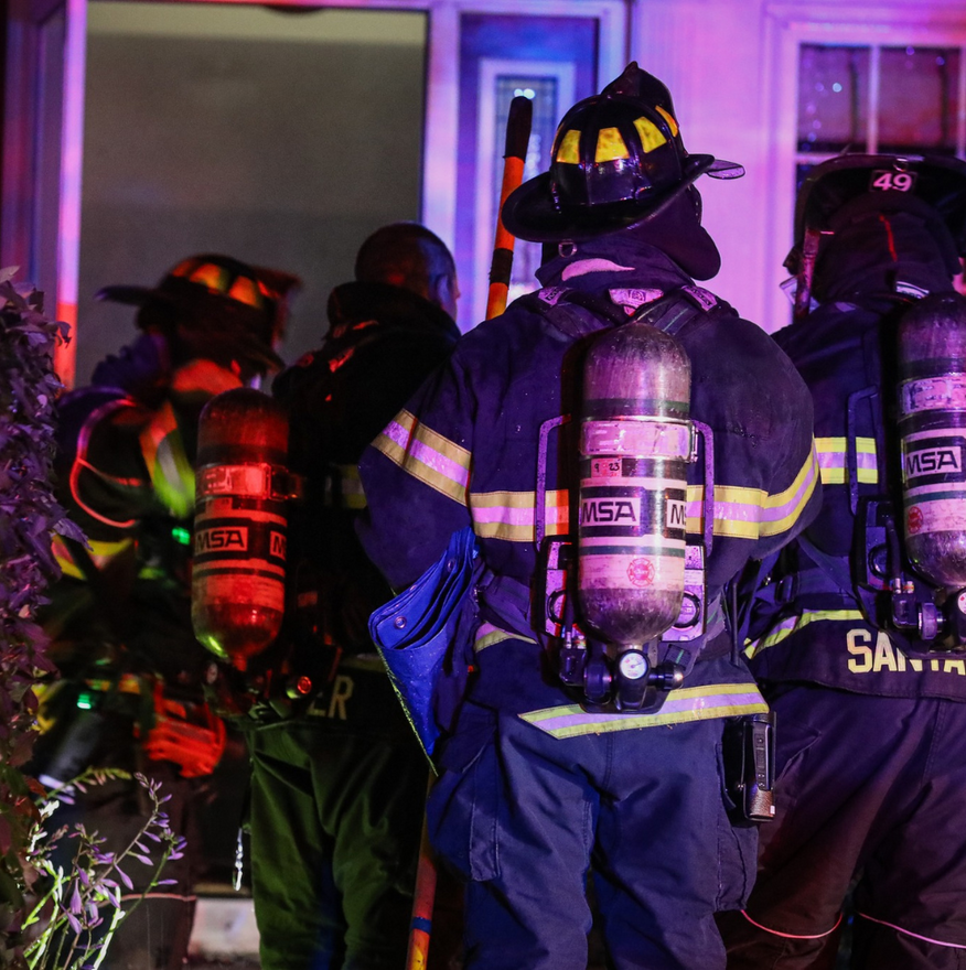 Firefighters in protective gear stand near a building entrance, with oxygen tanks on their backs.