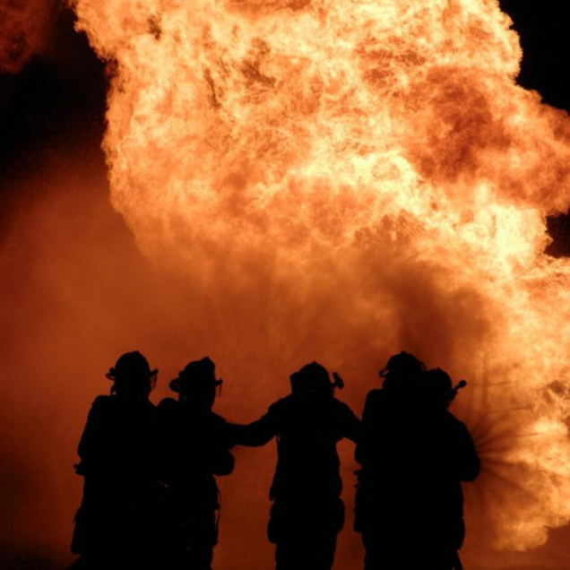 Firefighters stand silhouetted against a massive wall of fire at night.