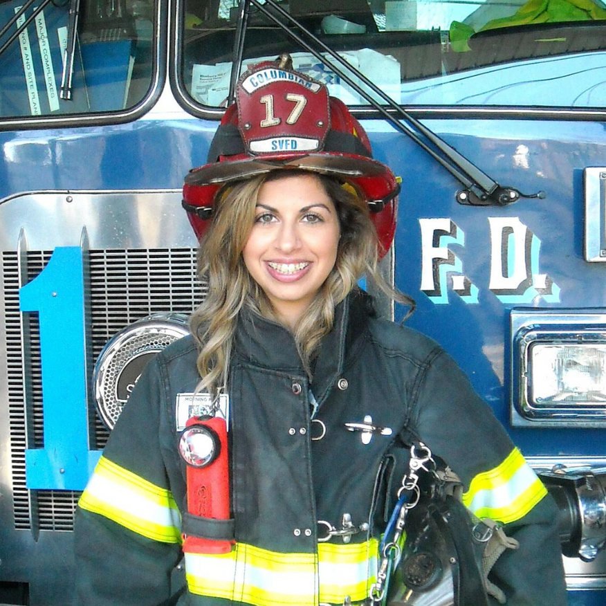 Firefighter in helmet and coat, smiling in front of a blue fire truck.
