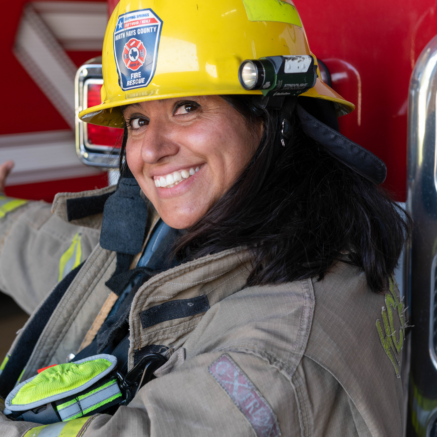 Firefighter smiling, wearing yellow helmet and protective gear, sitting near a fire truck.