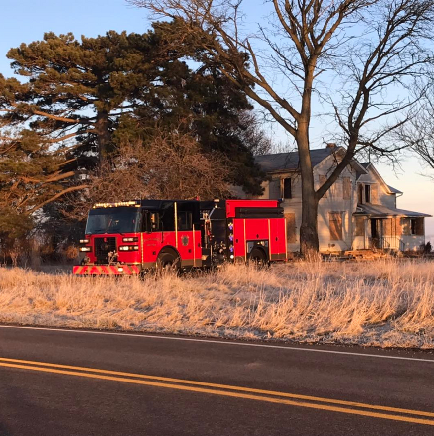 Red fire truck parked near an old, weathered house and tall trees on a roadside.
