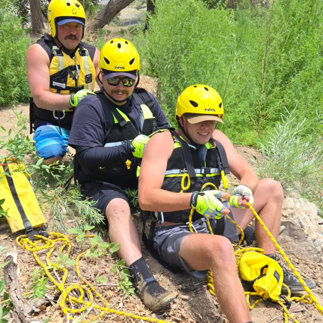Three people in yellow helmets and life vests practice roping near a waterway.