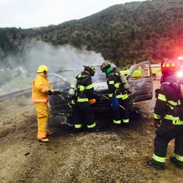 Firefighters extinguish a burning car on the side of a road.