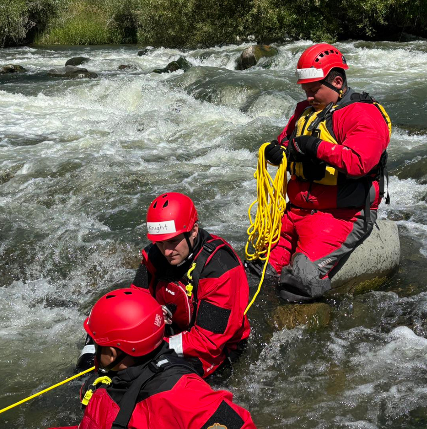 Three rescuers in red suits and helmets practice river rescue techniques in a whitewater stream.