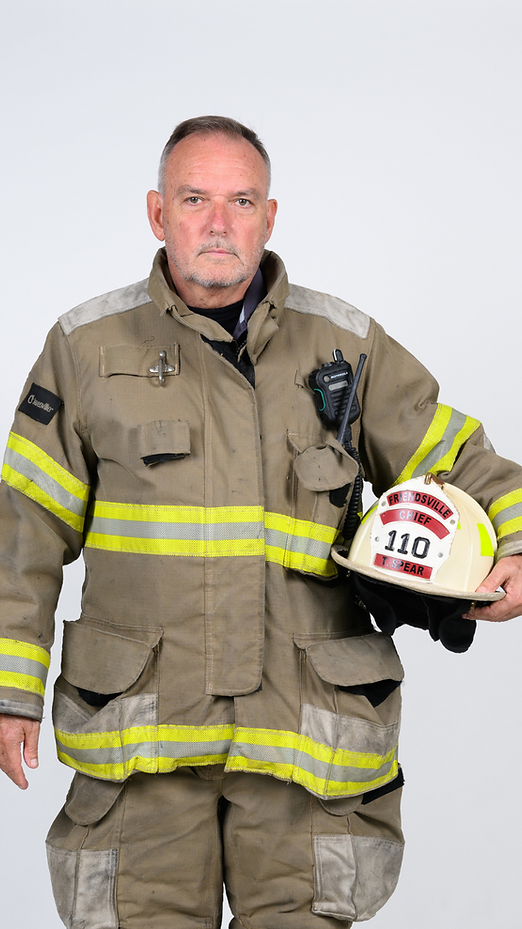 Firefighter holding helmet, wearing tan jacket with reflective stripes; facing camera, neutral expression.
