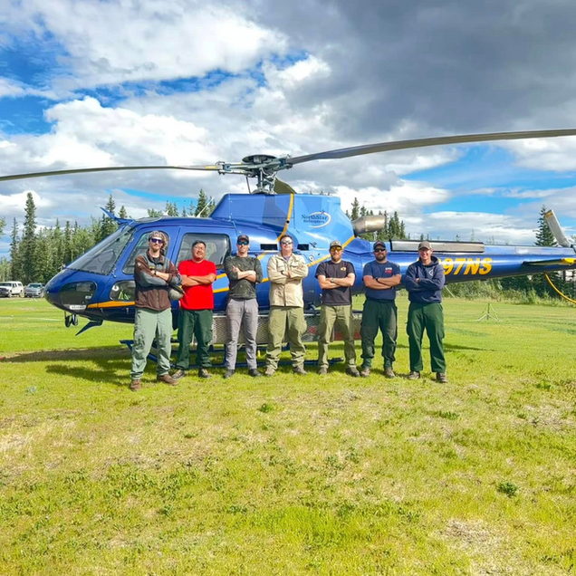 Firefighters pose in front of a blue helicopter on a grassy field under a cloudy sky.