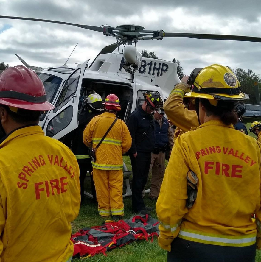 Firefighters at a helicopter, likely preparing for a medical transport. Yellow jackets, red helmets, cloudy day.