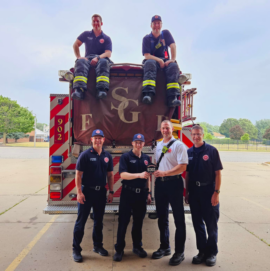Five firefighters pose with a fire truck. Two sit atop the truck; three stand in front.