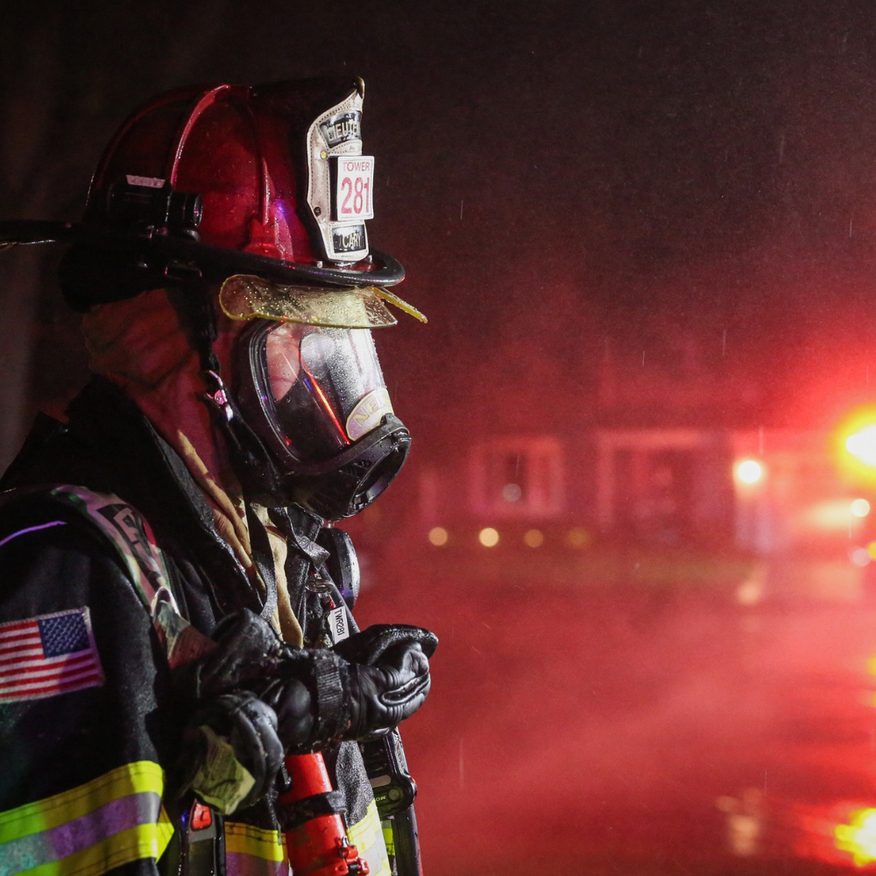 Firefighter in full gear, looking right, with red emergency lights in the background.