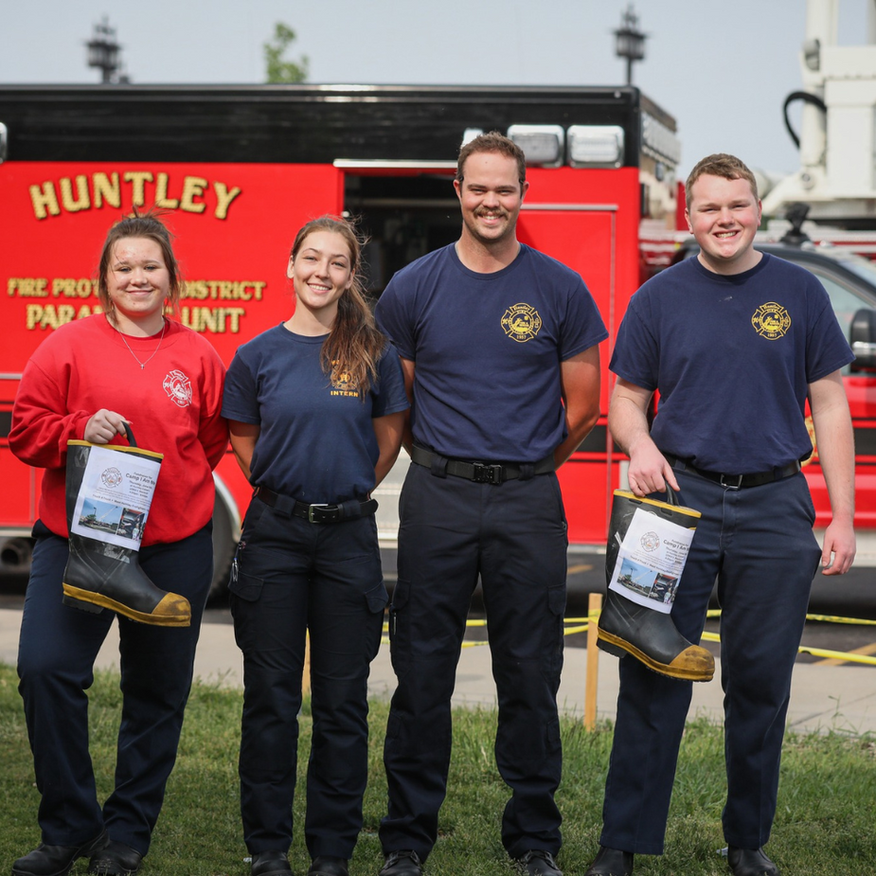 Four firefighters in uniform, holding boots in front of a fire truck; smiling outdoors.