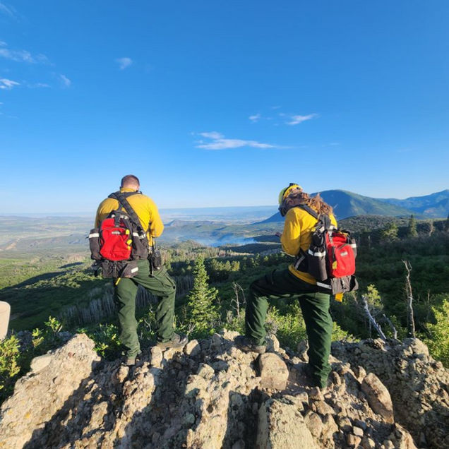 Two people in yellow fire gear survey a smoky valley from a rocky peak. Blue sky.