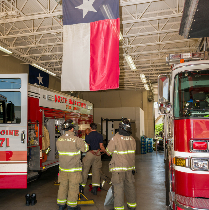 Firefighters in bunker gear in a fire station, Texas flag overhead, fire trucks in bays.