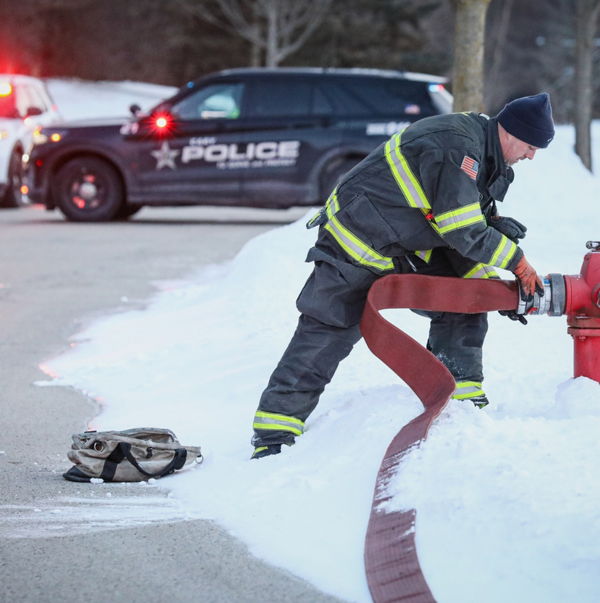 Firefighter connects a hose to a fire hydrant, snow surrounding them, police car in the background.