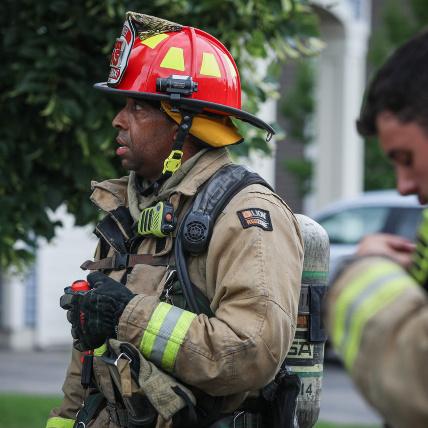 Firefighter in tan gear, red helmet, oxygen tank, looking intently.