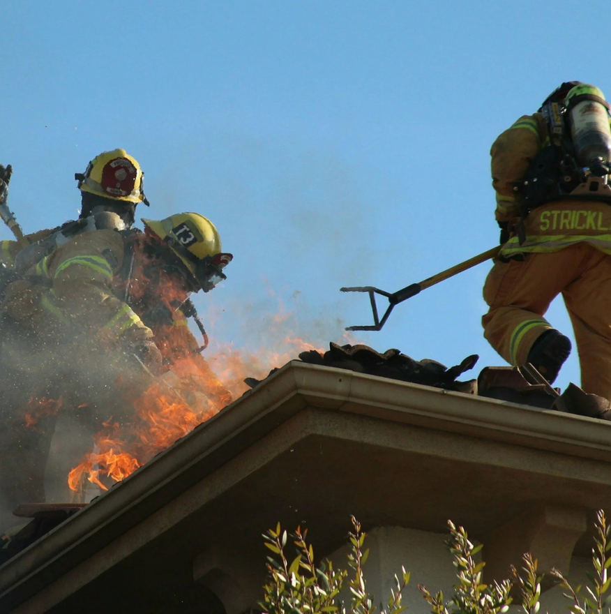 Firefighters on a roof battling a fire, with flames visible. One uses a tool, all wear protective gear.