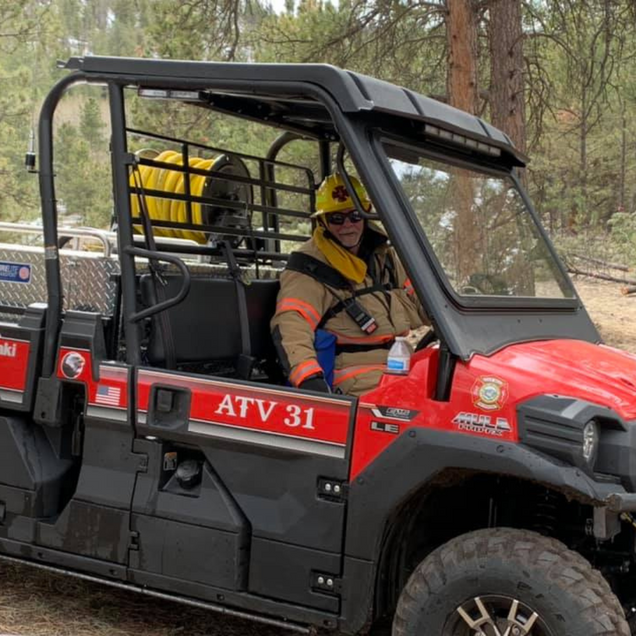 Firefighter in a red utility vehicle, wearing helmet and gear, in a wooded area.