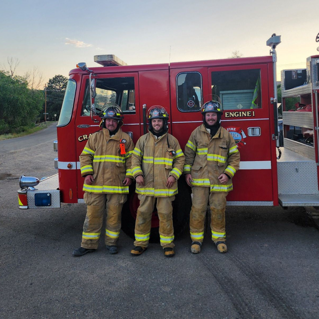 Three firefighters in gear standing in front of a red fire truck,