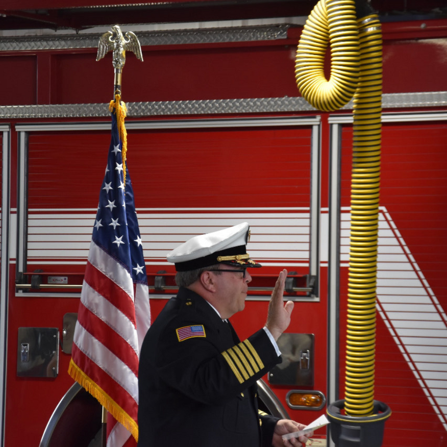 Fire chief taking an oath beside a fire truck, holding an American flag and a yellow hose.