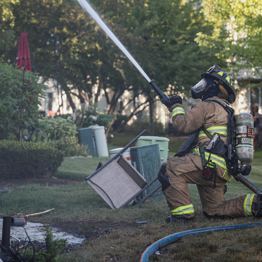 Firefighter kneels, spraying water from hose at a house fire in a yard; green grass, bushes, sky.