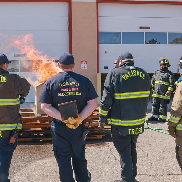 Firefighters watch a controlled burn outside a fire station. Flames erupt from a wooden structure.