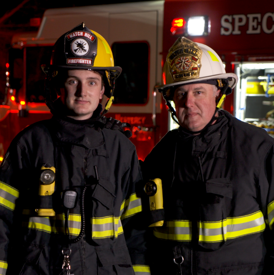 Two firefighters in uniform in front of a fire truck at night.