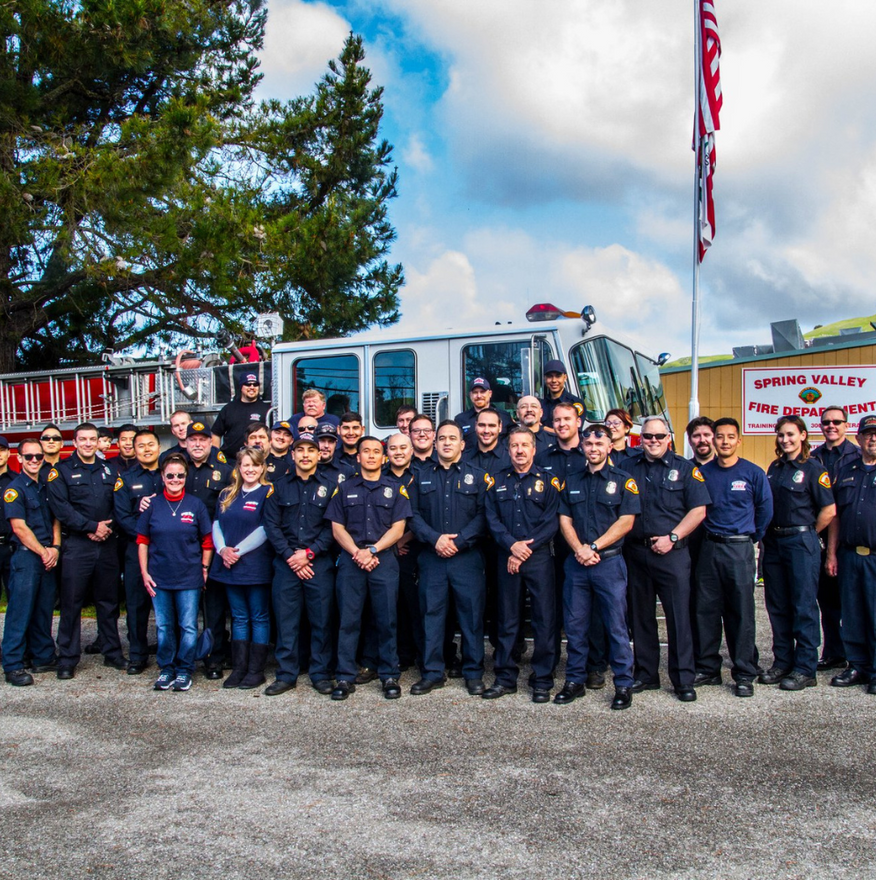 Firefighters pose in front of a fire engine. A sign indicates the fire department name.