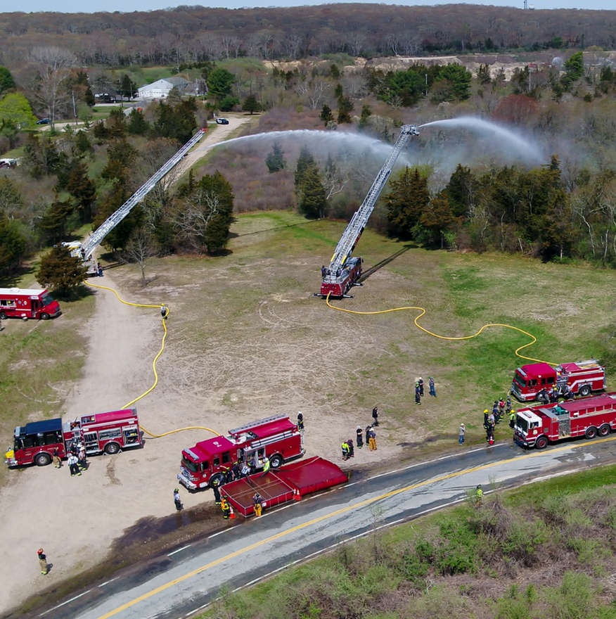 Fire trucks spraying water over a field; two extendable ladders, several vehicles, and responders are visible.