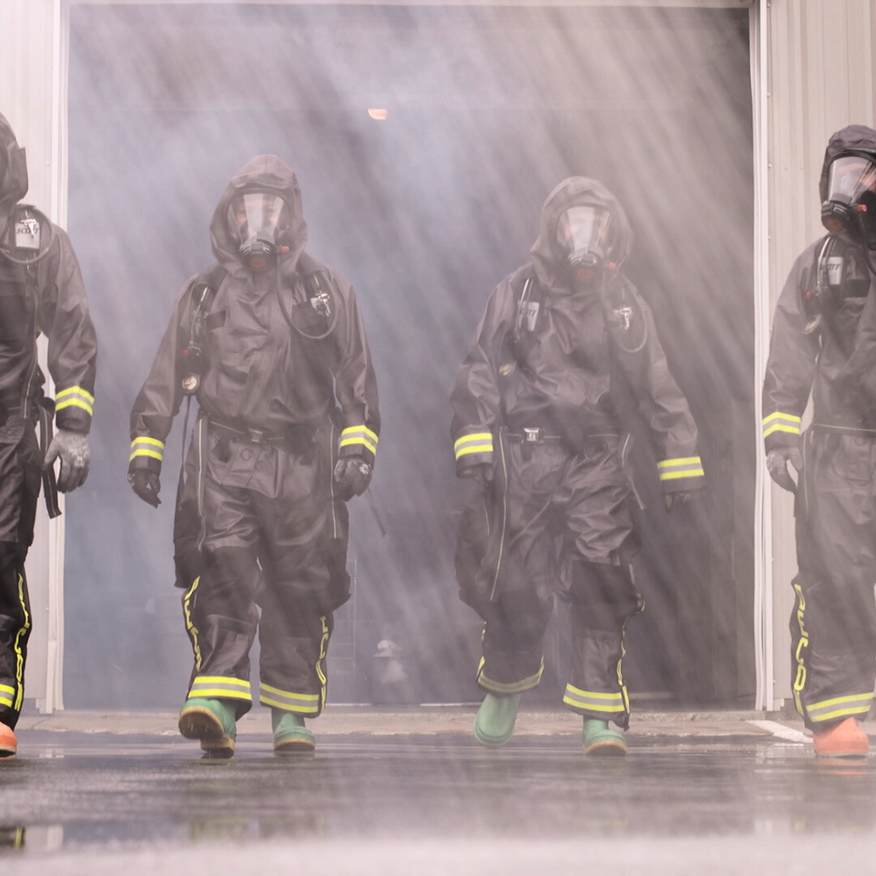 Four figures in hazmat suits emerge from smoky doorway. Yellow stripes on sleeves and boots.