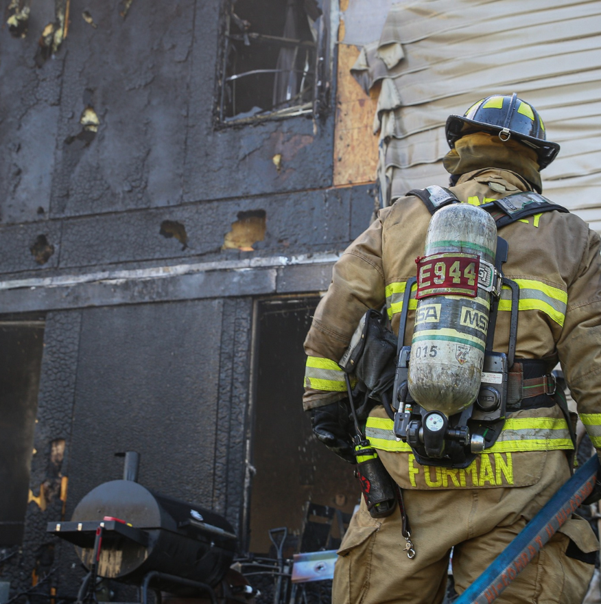 Firefighter in protective gear surveys a fire-damaged building. Blackened exterior, smoke visible.