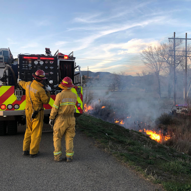 Firefighters in yellow suits, fire truck, battling a grass fire on roadside.