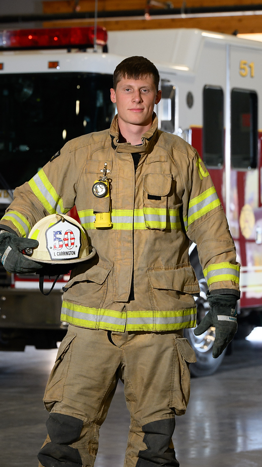 Firefighter in protective gear holding helmet in front of fire truck.