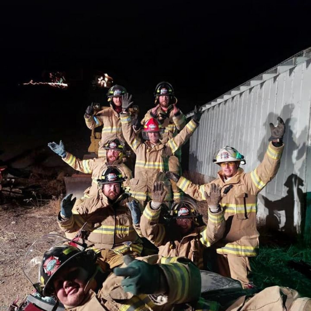 Firefighters in gear, arms raised, celebrating in front of a building at night.