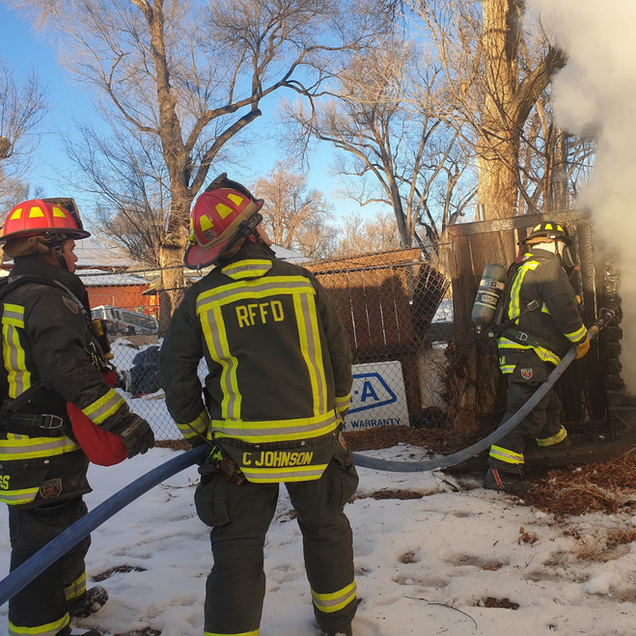Firefighters extinguishing a shed fire, wearing protective gear and using a hose on a snowy day.