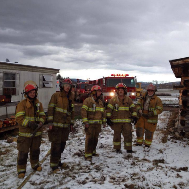 Firefighters in gear stand in snowy area with fire trucks and buildings.
