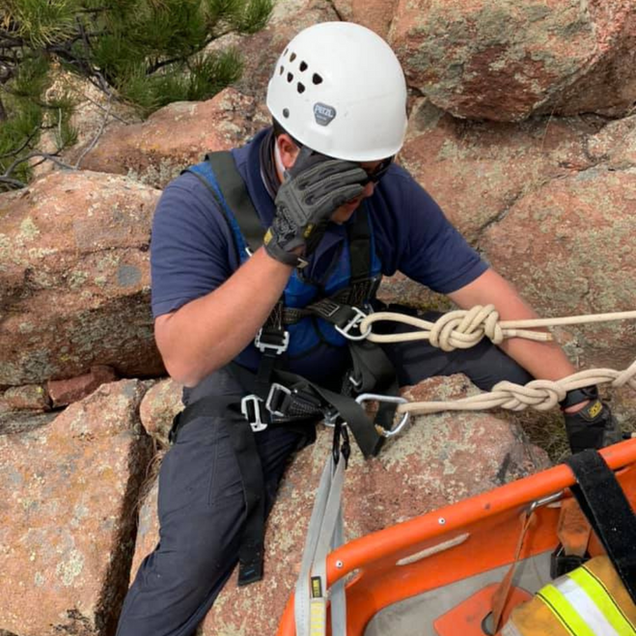 Rescue worker in harness on a rock face, adjusting sunglasses next to a rescue stretcher.