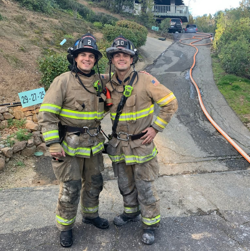 Two firefighters in gear pose on a sloped street with hoses, possibly after a fire.