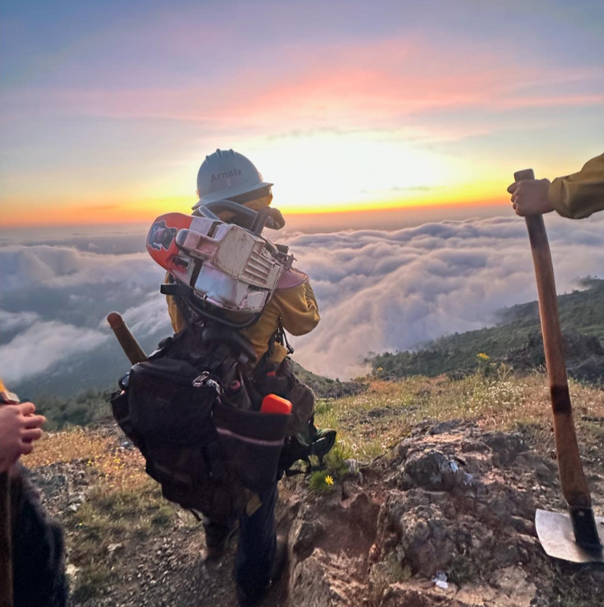 Firefighter on a mountain ridge, watching sunrise over clouds. Equipped with gear and shovel.