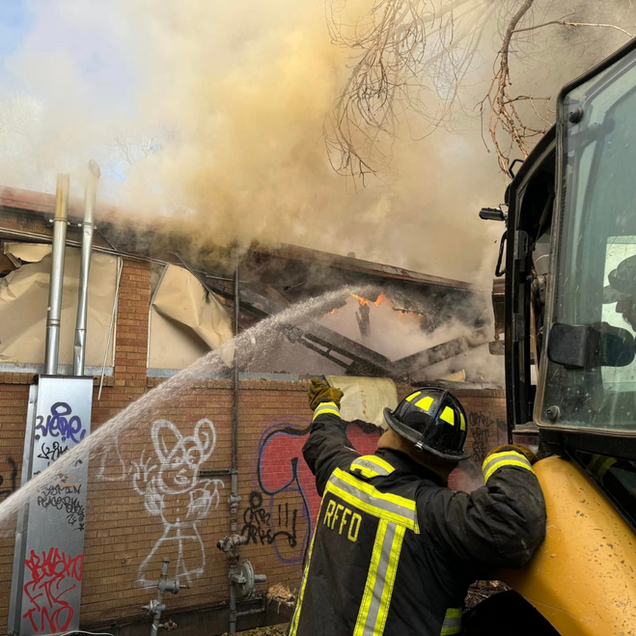 Firefighter spraying water on a burning building. Thick smoke billows as flames erupt from the roof.