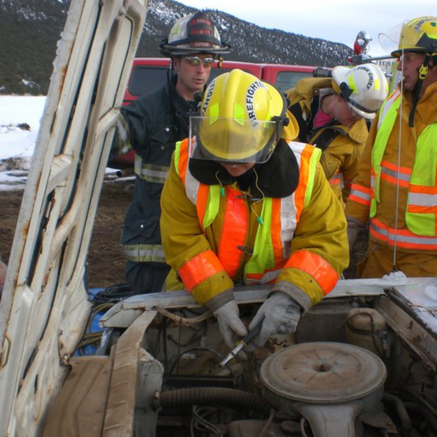 Firefighters examining a car engine in a snowy setting. One in a yellow coat is focused on the engine.