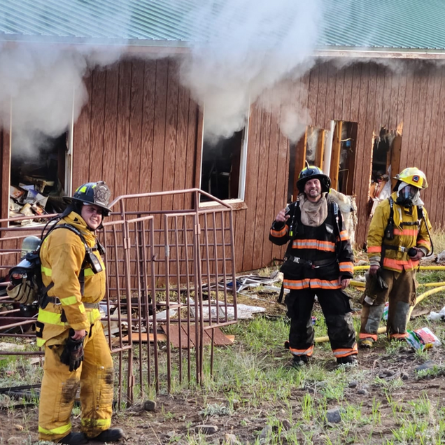 Firefighters at a burning building. Smoke billows from windows. One smiles, two wear breathing apparatus.