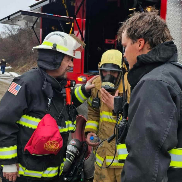 Firefighters near a fire truck, one in a respirator. Men are talking. Outdoor setting.