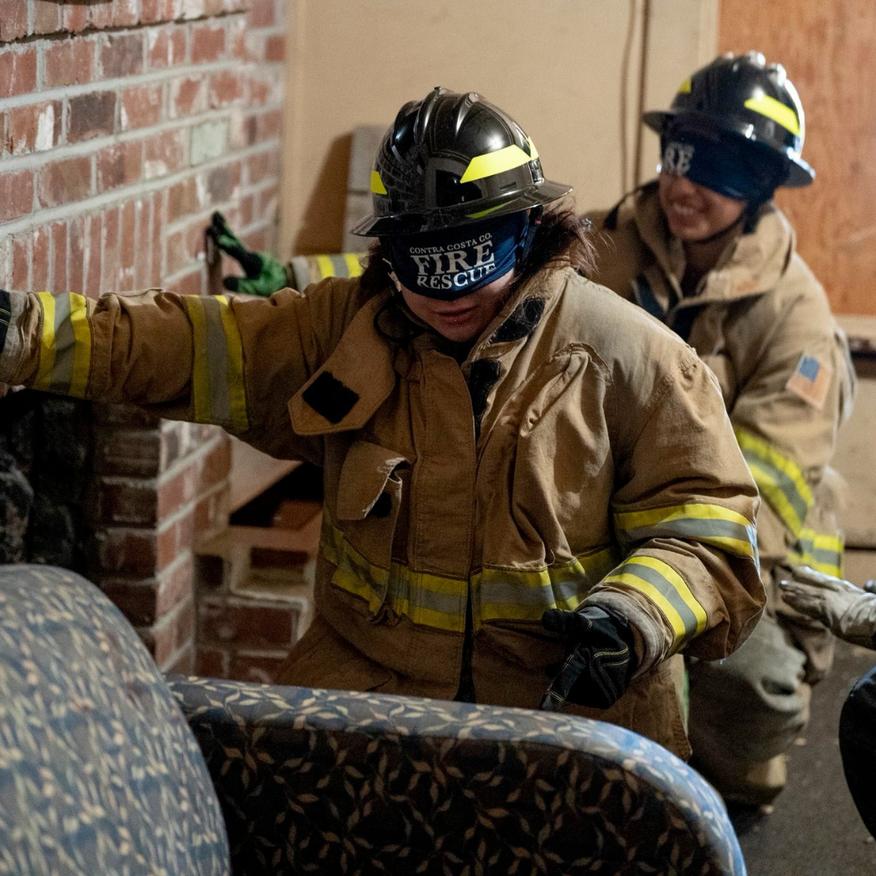 Firefighters in protective gear, one blindfolded, navigating a simulated structure, practicing search and rescue.