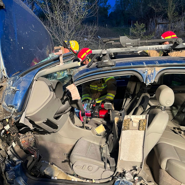 Firefighters at a vehicle collision. Damaged blue car with emergency personnel, red and yellow helmets. Evening setting.